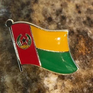 May include: A metal pin featuring the flag of Guinea-Bissau. The flag is red, yellow, and green with a star in the center of the red portion.