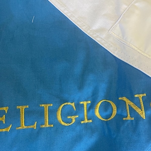 May include: A section of a flag with a light blue field and a white corner. The word "RELIGION" is embroidered in gold thread across the blue field. The flag is folded.