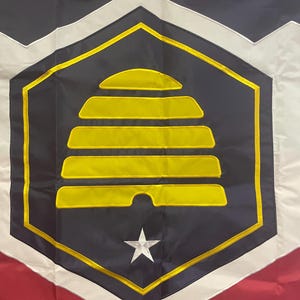 May include: A black and white flag with a yellow beehive design in the center. The beehive is surrounded by a gold border and a white star. The flag is made of fabric and has a textured surface.