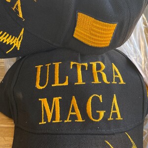 May include: Black baseball cap with gold embroidered text reading "ULTRA MAGA" and a gold embroidered American flag patch. The cap also features additional gold embroidery on the side.