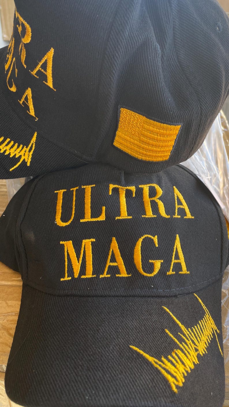 May include: Black baseball cap with gold embroidered text reading "ULTRA MAGA" and a gold embroidered American flag patch. The cap also features additional gold embroidery on the side.