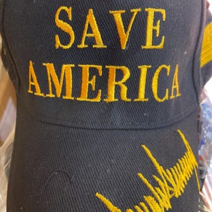 May include: Black baseball cap with gold embroidered text reading "SAVE AMERICA". The cap also features a gold embroidered signature on the brim. The hat is made of a durable fabric.