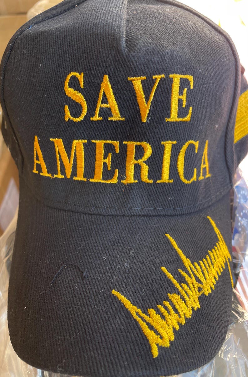 May include: Black baseball cap with gold embroidered text reading "SAVE AMERICA". The cap also features a gold embroidered signature on the brim. The hat is made of a durable fabric.