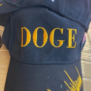 May include: Black baseball cap with the word "DOGE" embroidered in gold. The cap features an American flag patch and gold embroidery on the brim. The cap is made of a textured fabric.