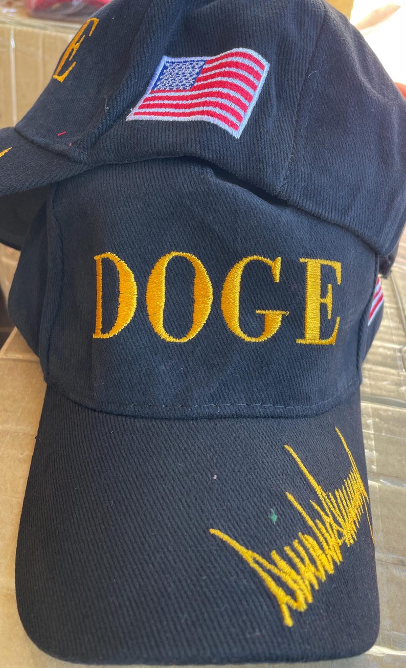 May include: Black baseball cap with the word "DOGE" embroidered in gold. The cap features an American flag patch and gold embroidery on the brim. The cap is made of a textured fabric.