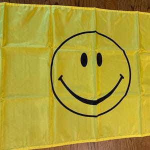 May include: A yellow flag with a large black smiley face design. The flag has a white reinforced edge with eyelets for hanging. The smiley face has two black oval eyes and a curved black mouth.