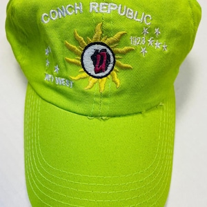 May include: A lime green baseball cap with white embroidery. The front of the cap reads "Conch Republic" with a sun and a "W" in a circle. The sun has 12 rays and the text "1928" is above the sun. There are five stars to the right of the sun and the text "To West" is below the sun.