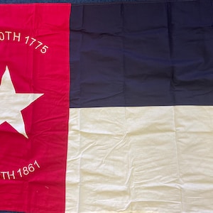 May include: A red, white, and blue flag with a single white star in the center. The flag has the text "MAY 20TH 1775" and "MAY 20TH 1861" printed on it.