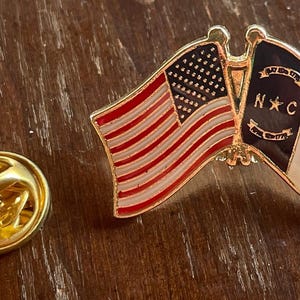 Puede incluir: Un pin dorado con la bandera estadounidense y la bandera de Carolina del Norte. La bandera estadounidense tiene rayas rojas y blancas y un campo de estrellas. La bandera de Carolina del Norte tiene un dise&ntilde;o rojo y blanco con las letras "N C".