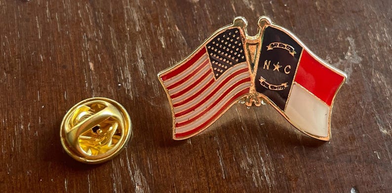 Puede incluir: Un pin dorado con la bandera estadounidense y la bandera de Carolina del Norte. La bandera estadounidense tiene rayas rojas y blancas y un campo de estrellas. La bandera de Carolina del Norte tiene un dise&ntilde;o rojo y blanco con las letras "N C".