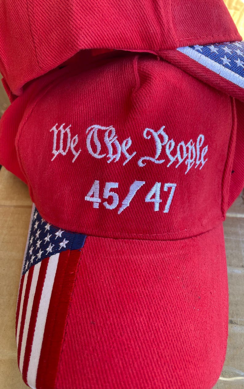 May include: Red baseball cap with white embroidered text "We The People" and the numbers "45/47". The cap features an American flag design on the brim and side. The cap is made of a textured fabric.