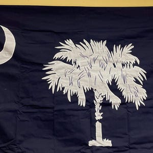 May include: A dark blue flag with a white crescent moon in the upper left corner and a white palmetto tree in the center. The tree is stylized with a single trunk and a large, bushy crown.