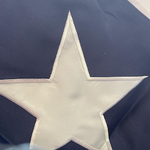 Puede incluir: Primer plano de una bandera azul marino con una gran estrella blanca en el centro. La estrella est&aacute; delineada con costuras blancas. La bandera tiene una apariencia texturizada.