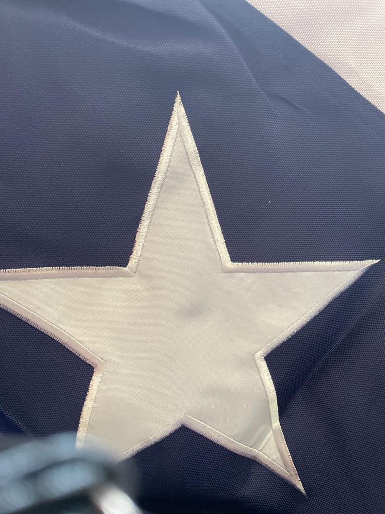Puede incluir: Primer plano de una bandera azul marino con una gran estrella blanca en el centro. La estrella est&aacute; delineada con costuras blancas. La bandera tiene una apariencia texturizada.