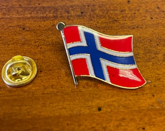 May include: A pin featuring the flag of Norway. The pin has a red background with a white-bordered blue cross. The pin is made of metal with a gold-colored backing and pin.