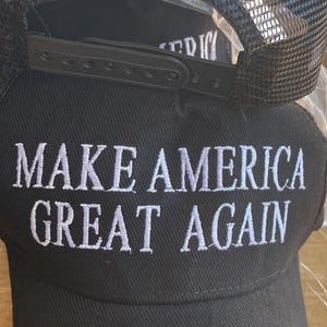 May include: Black baseball cap with white embroidered text reading "MAKE AMERICA GREAT AGAIN". The top of the cap has the word "TRUMP" embroidered in white. The cap features a black mesh back.