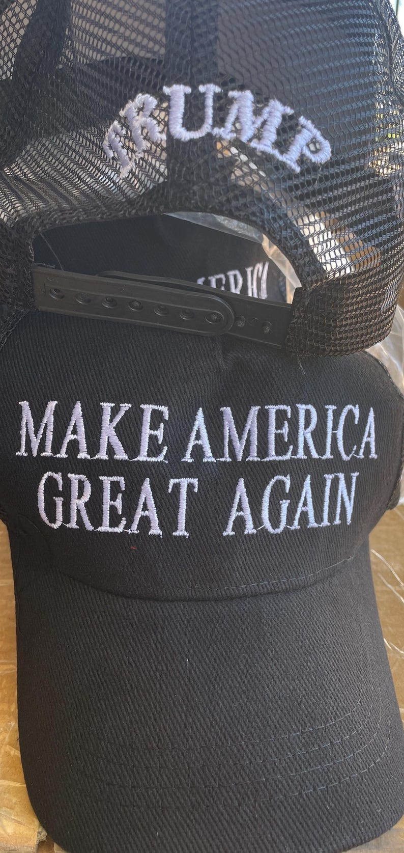 May include: Black baseball cap with white embroidered text reading "MAKE AMERICA GREAT AGAIN". The top of the cap has the word "TRUMP" embroidered in white. The cap features a black mesh back.