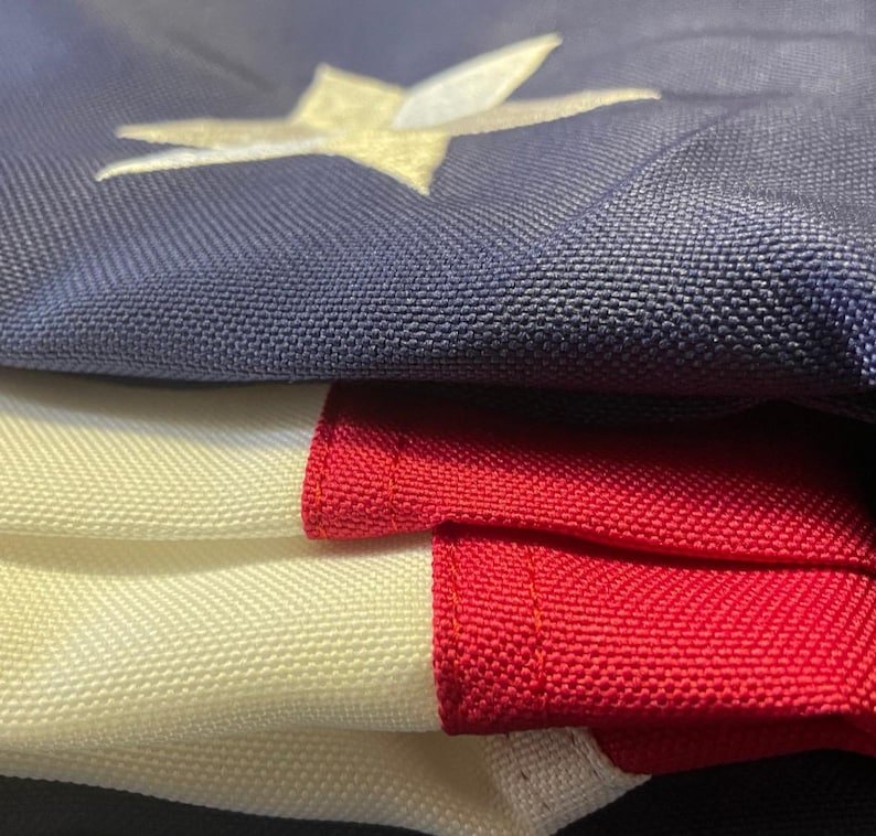 Puede incluir: Primer plano de una bandera estadounidense doblada. La bandera presenta un campo azul con una estrella dorada y rayas rojas y blancas. La tela tiene un tejido texturizado.