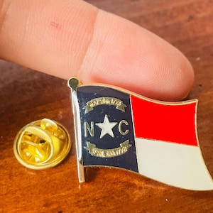 Puede incluir: Un pin dorado con el dise&ntilde;o de la bandera de Carolina del Norte. La bandera presenta una secci&oacute;n roja y blanca, una secci&oacute;n azul con una estrella y las letras "N" y "C", y los textos "May 20th 1775" y "April 12th 1776".