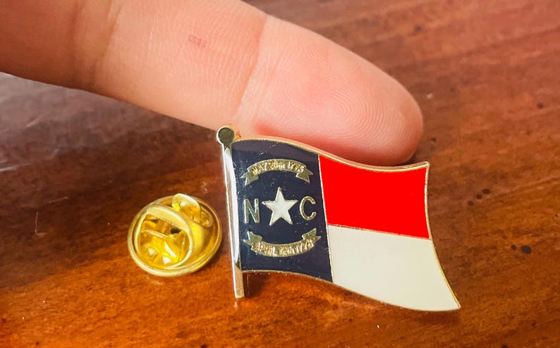 Puede incluir: Un pin dorado con el dise&ntilde;o de la bandera de Carolina del Norte. La bandera presenta una secci&oacute;n roja y blanca, una secci&oacute;n azul con una estrella y las letras "N" y "C", y los textos "May 20th 1775" y "April 12th 1776".