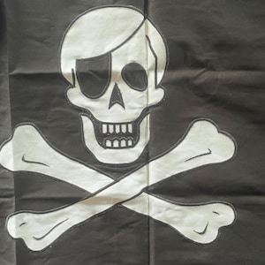 May include: A black flag featuring a large, white, embroidered skull and crossbones design. The skull has a prominent black eye patch over one eye, and the entire graphic is outlined with black stitching against the dark fabric background.