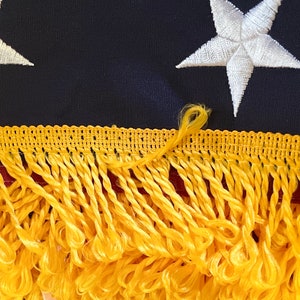 K&ouml;nnte beinhalten: Nahaufnahme einer goldenen Fransenborte auf einem marineblauen Stoff mit wei&szlig;en gestickten Sternen. Die Franse ist ein h&auml;ufiges dekoratives Element auf amerikanischen Flaggen.