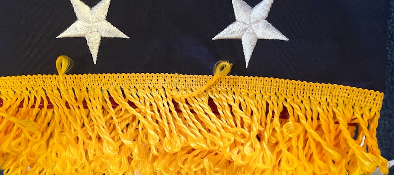 K&ouml;nnte beinhalten: Nahaufnahme einer goldenen Fransenborte auf einem marineblauen Stoff mit wei&szlig;en gestickten Sternen. Die Franse ist ein h&auml;ufiges dekoratives Element auf amerikanischen Flaggen.