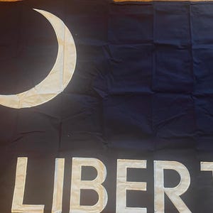 Puede incluir: Una bandera negra con una luna creciente blanca en la esquina superior izquierda y la palabra "LIBERTAD" en letras blancas grandes en la parte inferior. La bandera es de tela oscura.