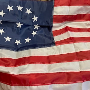 May include: A close-up of a 13-star American flag, a historical version of the flag with 13 white stars in a circle on a blue field, representing the original 13 colonies. The flag is made of red, white, and blue fabric and is slightly wrinkled.