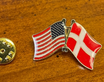 May include: A gold-colored pin with two flags, the American flag and the Danish flag, attached to a gold-colored pin back.