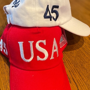 May include: Two baseball caps, one on top of the other. The top cap is white with the number "45" in navy blue and a navy blue emblem. The bottom cap is red with the word "USA" in white.