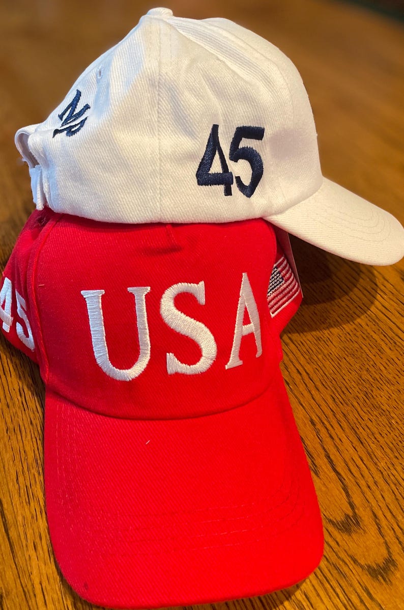 May include: Two baseball caps, one on top of the other. The top cap is white with the number "45" in navy blue and a navy blue emblem. The bottom cap is red with the word "USA" in white.