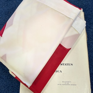 May include: A folded white and red flag with the text "UNITED STATES OF AMERICA" and "BY AUTHORITY OF THE UNITED CONFEDERATE VETERANS" printed on the white portion of the flag.