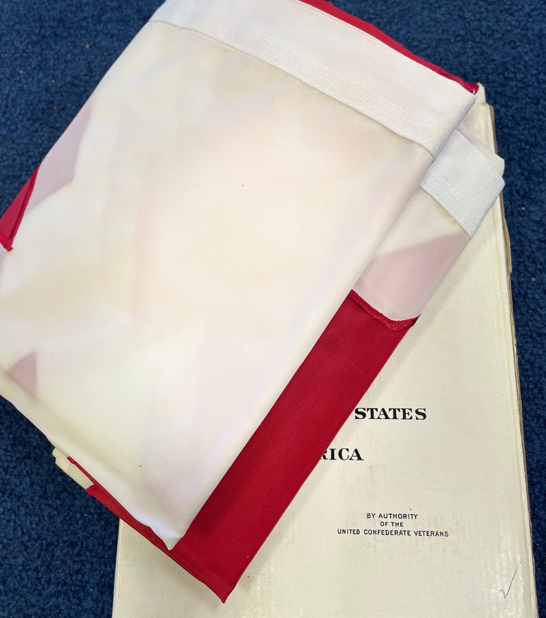 May include: A folded white and red flag with the text "UNITED STATES OF AMERICA" and "BY AUTHORITY OF THE UNITED CONFEDERATE VETERANS" printed on the white portion of the flag.