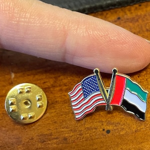 May include: A gold pin with two crossed flags. The flag on the left is the American flag, and the flag on the right is the flag of the United Arab Emirates.