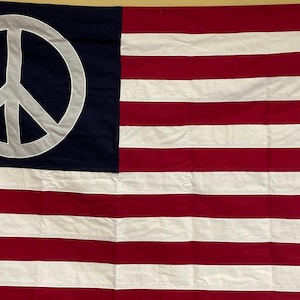 May include: A rectangular flag featuring a dark blue canton in the upper left corner with a large, light gray peace symbol centered within it. The remainder of the flag consists of alternating horizontal red and white stripes across the field.