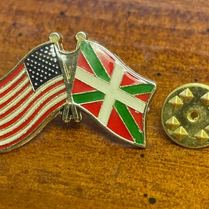 以下が含まれることがあります： 2つの旗が交差した金色のピン。旗はアメリカの国旗とバスク地方の旗です。ピンは円形のデザインが施された金色の裏側を持っています。