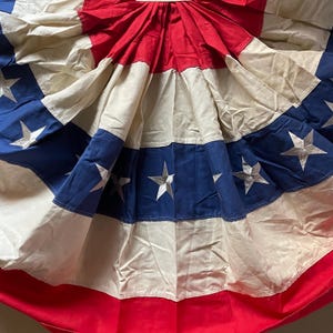 May include: A pleated fabric bunting featuring a patriotic design with horizontal bands of red, off-white, and blue. The blue section is adorned with white five-pointed stars arranged in a row, creating a decorative fan-shaped display.