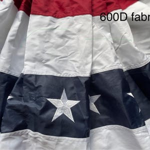 Puede incluir: Primer plano de una tela con un dise&ntilde;o patri&oacute;tico. La tela presenta rayas rojas, blancas y azul marino y estrellas blancas bordadas. El texto "600D fabric" es visible en la tela.