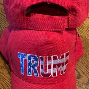 May include: Two red baseball caps, one on top of the other. The front cap features the word "TRUMP" embroidered in white and blue, with the letters "T" and "P" in red. The caps are made of a textured material.