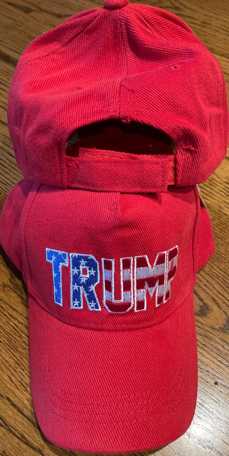 May include: Two red baseball caps, one on top of the other. The front cap features the word "TRUMP" embroidered in white and blue, with the letters "T" and "P" in red. The caps are made of a textured material.
