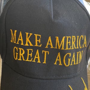 May include: Black baseball cap with the embroidered phrase "MAKE AMERICA GREAT AGAIN" in gold thread. The cap also features a gold embroidered signature. A small American flag is visible.
