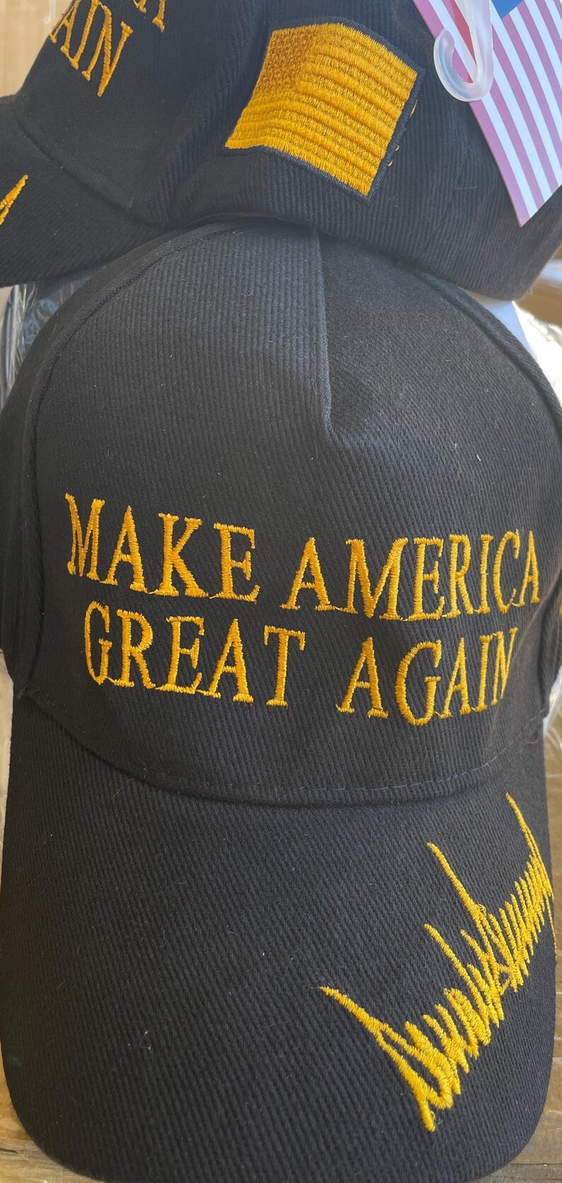 May include: Black baseball cap with the embroidered phrase "MAKE AMERICA GREAT AGAIN" in gold thread. The cap also features a gold embroidered signature. A small American flag is visible.