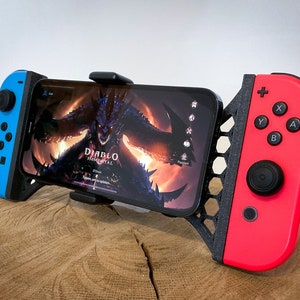 Pode incluir: Um controlador Nintendo Switch preto e vermelho com um suporte para telefone no meio. O telefone está exibindo o jogo Diablo Immortal.