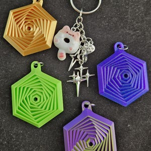 May include: Four hexagonal keychains with a swirling pattern in different colors: gold, green, purple, and a gradient of purple and green. The gold keychain has a small charm attached to it with a cartoon animal and a heart that says "Made with love".