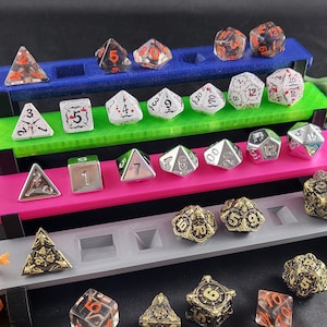 May include: A tiered acrylic display stand for dice, featuring three levels in blue, green, and pink. The stand is filled with various dice in different colors and styles, including clear, white, black, and silver.