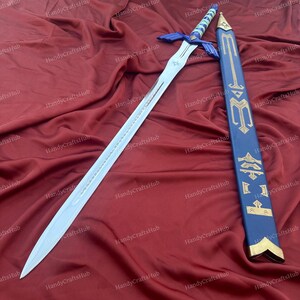 Puede incluir: Una espada plateada con empu&ntilde;adura azul y blanca y una vaina azul a juego con detalles dorados. La espada est&aacute; sobre una tela roja. La vaina presenta s&iacute;mbolos dorados.