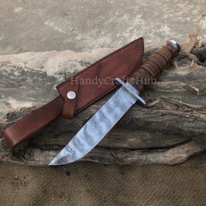 Może przedstawiać: N&oacute;ż myśliwski ze stali damasceńskiej z brązową sk&oacute;rzaną rękojeścią i pochwą. Ostrze ma unikalny wz&oacute;r, a rękojeść jest owinięta sk&oacute;rą. Pochwa ma zapięcie na zatrzask. Widoczny jest tekst "HandyCraftsHub".
