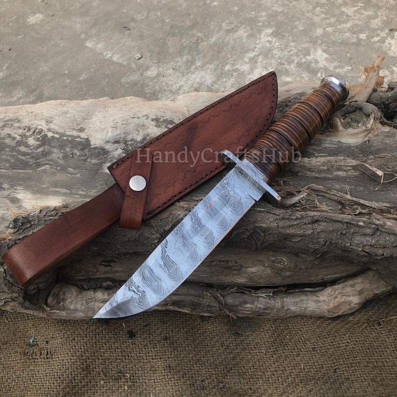 Może przedstawiać: N&oacute;ż myśliwski ze stali damasceńskiej z brązową sk&oacute;rzaną rękojeścią i pochwą. Ostrze ma unikalny wz&oacute;r, a rękojeść jest owinięta sk&oacute;rą. Pochwa ma zapięcie na zatrzask. Widoczny jest tekst "HandyCraftsHub".