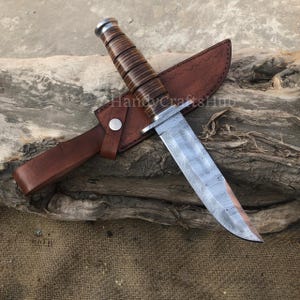 Może przedstawiać: N&oacute;ż myśliwski ze stali damasceńskiej z brązową sk&oacute;rzaną rękojeścią i pochwą. Ostrze ma unikalny wz&oacute;r, a rękojeść wykonana jest ze sk&oacute;rzanych warstw. Pochwa jest zabezpieczona metalowym zatrzaskiem. N&oacute;ż spoczywa na kawałku drewna.